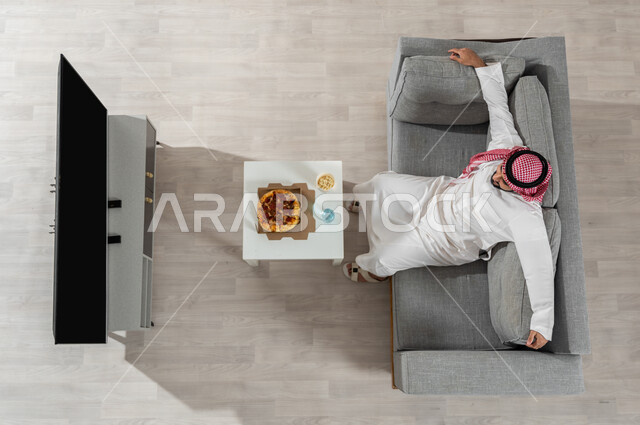 Spending leisure time watching TV programmes, a vertical overhead portrait of a Saudi Gulf Arab man wearing a traditional dress with a shemagh sitting on the sofa in the living room, gestures of rest and relaxation, displaying a blank black screen on the TV, beige background