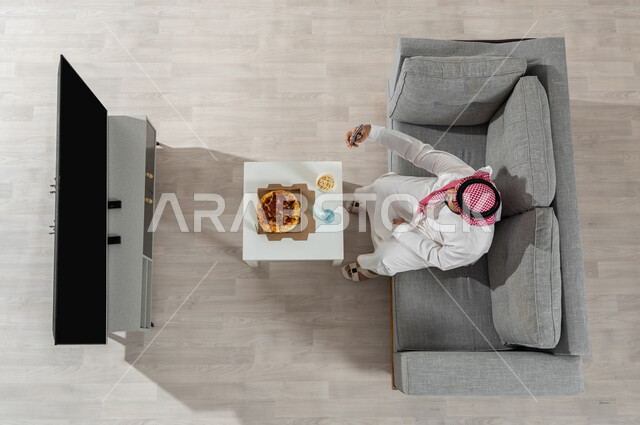 التقاط صورة سيلفي للذكرى , بورتريه علوي عامودي لرجل عربي خليجي سعودي يرتدي الزي التقليدي مع الشماغ يجلس على الأريكة في غرفة المعيشة يمسك الهاتف المحمول بيده , وقت تناول الغداء , عرض شاشة سوداء فارغة عبر التلفاز, خلفية بيج