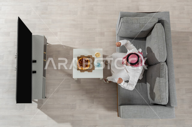 الاستمتاع بمشاهدة البرامج التلفزيونية أثناء تناول الغداء ,بورتريه علوي عامودي لرجل عربي خليجي سعودي يرتدي الزي التقليدي مع الشماغ يجلس على الأريكة في غرفة المعيشة , إيماءات التفاعل و الحماس, عرض شاشة سوداء فارغة عبر التلفاز, خلفية بيج