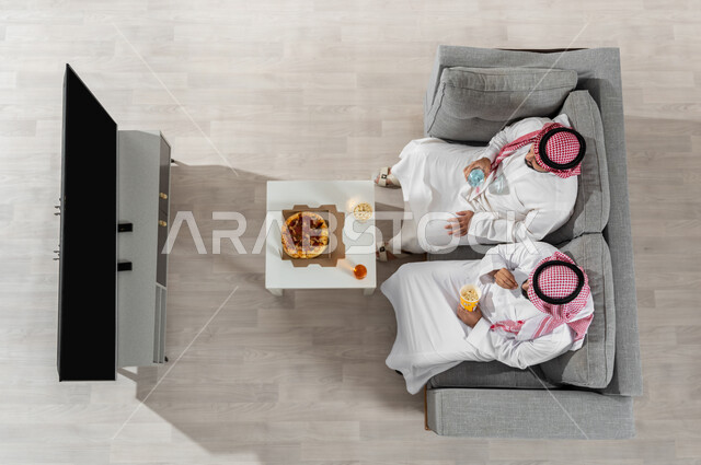 مشاركة الأصدقاء الأوقات الممتعة , بورتريه علوي عامودي لرجلان عربيان خليجيان سعوديان يرتديان الزي التقليدي يجلسان على الأريكة في غرفة المعيشة , تناول الغداء أثناء مشاهدة البرامج التلفزيونية , عرض شاشة سوداء عبر التلفاز , خلفية بيج