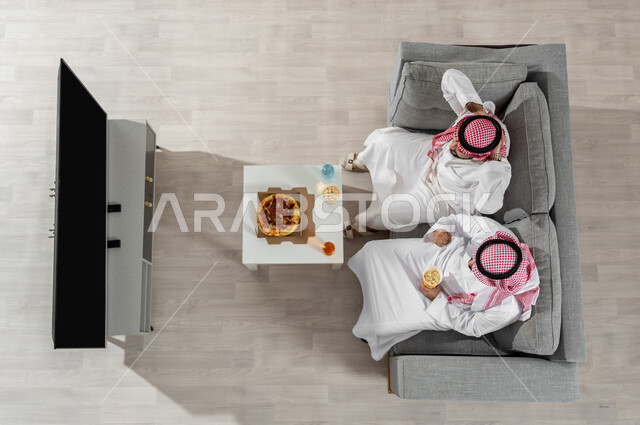 Watching TV programs while eating lunch, vertical portrait of two Saudi Gulf Arab men wearing traditional clothing with a shemagh, sitting on the sofa in the living room eating, black screen displayed on the TV, beige background