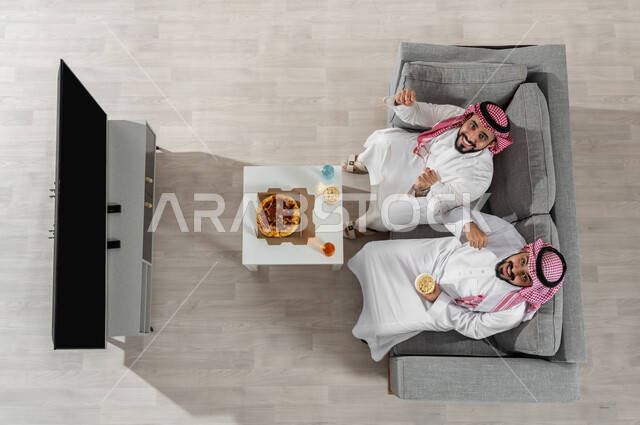 Looking at the camera with gestures of happiness and pleasure, friends sharing lunch, a vertical overhead portrait of two Saudi Gulf Arab men wearing traditional clothing with a shemagh, sitting on the sofa in the living room eating pizza, a black screen shown on the TV, beige background