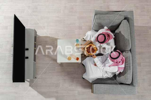 قضاء وقت الغداء مع الأصدقاء , بورتريه علوي عامودي لرجلان عربيان خليجيان سعوديان يرتديان الزي التقليدي مع الشماغ يجلسان على الأريكة في غرفة المعيشة يقومان بتناول البيتزا , عرض شاشة سوداء عبر التلفاز , خلفية بيج