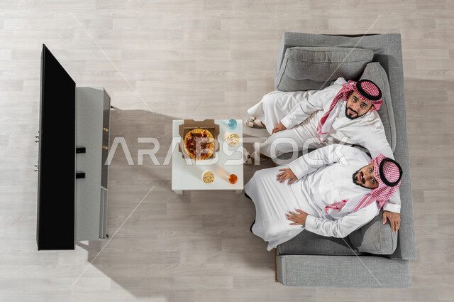 مشاركة وقت الغداء مع الأصدقاء، بورتريه علوي عامودي لشابان عربيان خليجيان سعوديان يرتديان الزي التقليدي يجلسان على الأريكة في غرفة المعيشة، النظر إلى الكاميرا بإيماءات السعادة والراحة، عرض شاشة التلفاز السوداء الفارغة، خلفية بيج