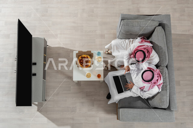 مشاركة الأوقات مع الأصدقاء، بورتريه علوي عامودي لشابان عربيان خليجيان سعوديان يرتديان الزي التقليدي يجلسان على الأريكة في غرفة المعيشة ويقومان بتناول البيتزا، استخدام الأجهزة الإلكترونية، عرض شاشة التلفاز السوداء الفارغة، خلفية بيج