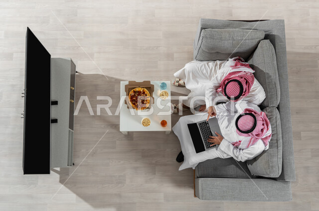 مشاركة الأوقات مع الأصدقاء، بورتريه علوي عامودي لشابان عربيان خليجيان سعوديان يرتديان الزي التقليدي يجلسان على الأريكة في غرفة المعيشة ويقومان بتناول البيتزا، استخدام الأجهزة الإلكترونية، عرض شاشة التلفاز السوداء الفارغة، خلفية بيج