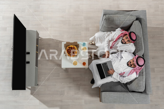مشاركة الأوقات مع الأصدقاء، بورتريه علوي عامودي لشابان عربيان خليجيان سعوديان يرتديان الزي التقليدي يجلسان على الأريكة في غرفة المعيشة ويقومان بتناول البيتزا، النظر إلى الكاميرا بإيماءات السعادة، استخدام الأجهزة الإلكترونية، عرض شاشة التلفاز الفارغة، خلفية بيج
