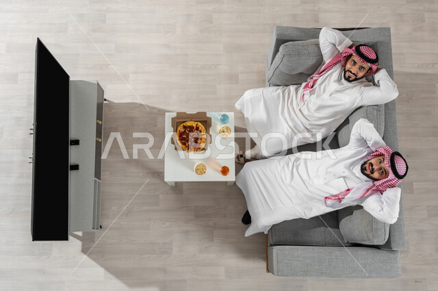 مشاركة وقت الغداء مع الأصدقاء، بورتريه علوي عامودي لشابان عربيان خليجيان سعوديان يرتديان الزي التقليدي يجلسان على الأريكة في غرفة المعيشة ويضعون أيديهم خلف رأسهم، النظر إلى الكاميرا بإيماءات السعادة والراحة، عرض شاشة التلفاز السوداء الفارغة، خلفية بيج