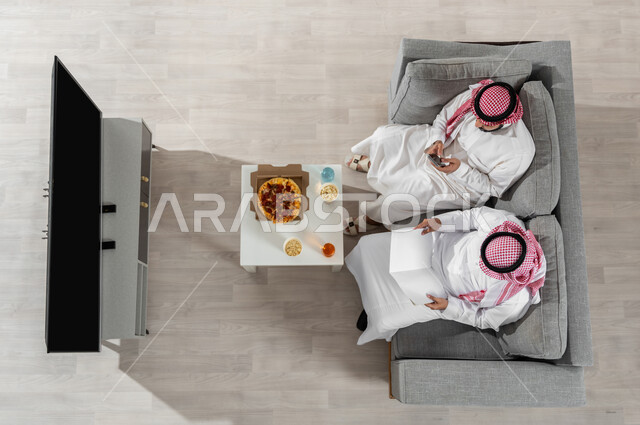 مشاركة وقت الغداء مع الأصدقاء، بورتريه علوي عامودي لشابان عربيان خليجيان سعوديان يرتديان الزي التقليدي يجلسان على الأريكة في غرفة المعيشة ويقومان بأنشطة مختلفة، استخدام الأجهزة الإلكترونية، عرض شاشة التلفاز السوداء الفارغة، خلفية بيج