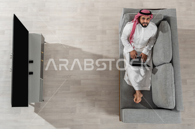 قضاء الوقت بإنجاز المهام المطلوبة، بورتريه علوي عامودي لشاب عربي خليجي سعودي يرتدي الزي التقليدي ينظر إلى الكاميرا ويجلس على الأريكة في غرفة المعيشة ويقوم بالعمل من خلال جهاز الحاسوب المحمول، عرض شاشة التلفاز السوداء الفارغة، خلفية بيج