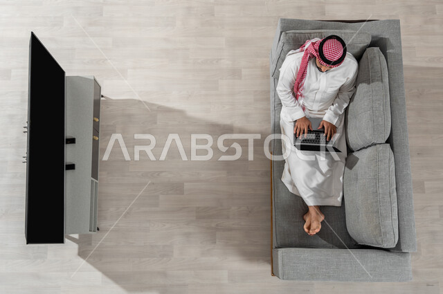 قضاء الوقت بإنجاز المهام المطلوبة، بورتريه علوي عامودي لشاب عربي خليجي سعودي يرتدي الزي التقليدي ينظر إلى الكاميرا ويجلس على الأريكة في غرفة المعيشة ويقوم بالعمل من خلال جهاز الحاسوب المحمول، عرض شاشة التلفاز السوداء الفارغة، خلفية بيج