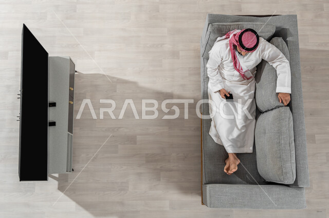 استعمال الأجهزة الالكترونية، قضاء وقت الاستراحة في المنزل، بورتريه لرجل عربي خليجي سعودي يرتدي الزي التقليدي يجلس على الأريكة في غرفة المعيشة ويقوم باستخدام الهاتف الذكي، عرض شاشة التلفاز والجوال سوداء فارغة، خلفية بيج