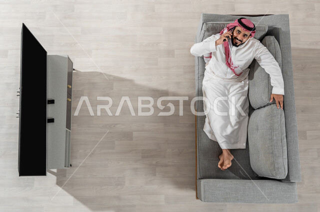 القيام بمكالمة هاتفية باستعمال الأجهزة الالكترونية، قضاء وقت الاستراحة في المنزل، بورتريه لرجل عربي خليجي سعودي يرتدي الزي التقليدي يجلس على الأريكة في غرفة المعيشة ويقوم باستخدام الهاتف الذكي، النظر إلى الكاميرا بإيماءات البهجة والسعادة، عرض شاشة التلفاز سوداء فارغة، خلفية بيج