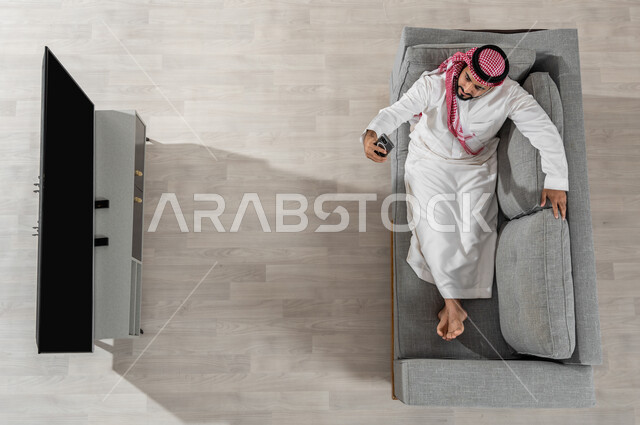 الاستفادة من الأجهزة الالكترونية، قضاء وقت الاستراحة في المنزل، بورتريه لرجل عربي خليجي سعودي يرتدي الزي التقليدي يجلس على الأريكة في غرفة المعيشة ويقوم باستخدام الهاتف الذكي والتقاط صور السيلفي، عرض شاشة سوداء فارغة، خلفية بيج