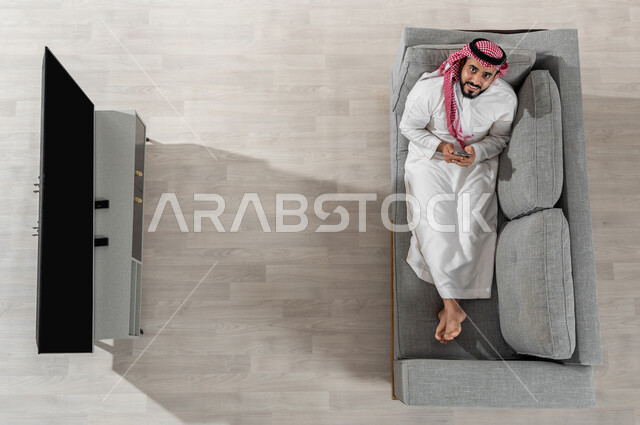 استعمال الأجهزة الالكترونية، قضاء وقت الاستراحة في المنزل، بورتريه لرجل عربي خليجي سعودي يرتدي الزي التقليدي يجلس على الأريكة في غرفة المعيشة ويقوم باستخدام الهاتف الذكي، النظر إلى الكاميرا بإيماءات البهجة والسعادة، عرض شاشة التلفاز السوداء الفارغة، خلفية بيج