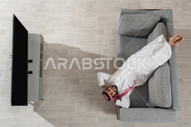 استعمال الأجهزة الالكترونية، الاسترخاء وقضاء وقت الاستراحة في المنزل، بورتريه لرجل عربي خليجي سعودي يرتدي الزي التقليدي يستلقي على الأريكة في غرفة المعيشة ويضع يديه خلف رأسه، النظر إلى الكاميرا بإيماءات السعادة والراحة، عرض شاشة سوداء فارغة، خلفية بيج