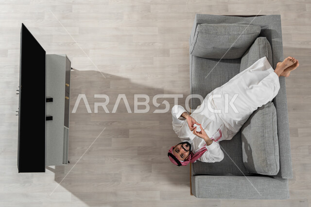 قضاء أوقات الاستراحة في المنزل، بورتريه لرجل عربي خليجي سعودي يرتدي الزي التقليدي يستلقي على الأريكة في غرفة المعيشة، النظر إلى الكاميرا بإيماءات السعادة والحب، استعمال الأجهزة الالكترونية، عرض شاشة سوداء فارغة، خلفية بيج