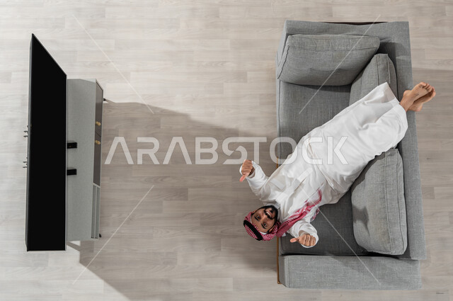 قضاء أوقات الاستراحة في المنزل، بورتريه لرجل عربي خليجي سعودي يرتدي الزي التقليدي يستلقي على الأريكة في غرفة المعيشة، النظر إلى الكاميرا بإيماءات السعادة والاعجاب، استعمال الأجهزة الالكترونية، عرض شاشة سوداء فارغة، خلفية بيج