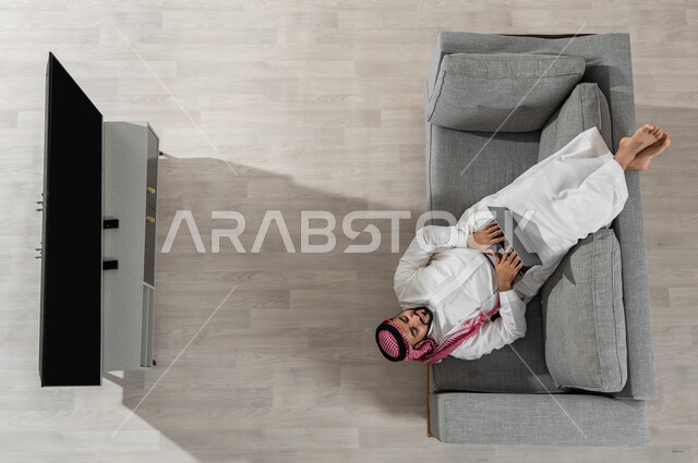قضاء الوقت في المنزل واستعمال الأجهزة الالكترونية، بورتريه علوي عامودي لشاب عربي خليجي سعودي يرتدي الزي التقليدي يستلقي على الأريكة في غرفة المعيشة ويقوم بالعمل على جهاز الحاسوب المحمول، عرض شاشة التلفاز السوداء الفارغة، خلفية بيج
