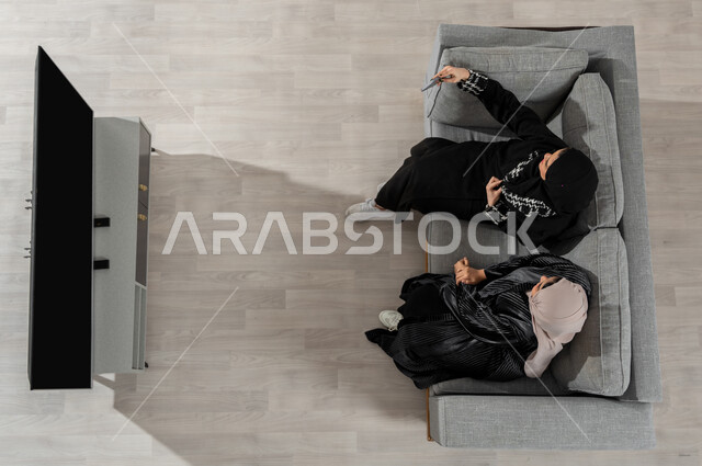 قضاء وقت ممتع في المنزل مع الأصدقاء، بورتريه علوي عامودي لفتاتان عربيتان خليجيتان سعوديتان ترتديان الحجاب والعباءة السوداء تجلسان على الأريكة في غرفة المعيشة، التقاط صور السيلفي للذكرى عرض شاشة الجوال واللابتوب السوداء الفارغة، خلفية بيج