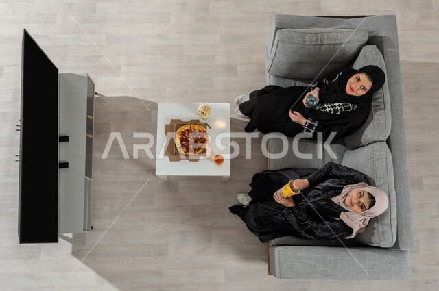 مشاركة الأوقات في المنزل مع الأصدقاء، بورتريه علوي عامودي لفتاتان عربيتان خليجيتان سعوديتان ترتديان الحجاب والعباءة السوداء تجلسان على الأريكة في غرفة المعيشة تقومان بتناول الفوشار وشرب الماء، النظر إلى الكاميرا بإيماءات السعادة والسرور، عرض شاشة التلفاز الفارغة، خلفية بيج