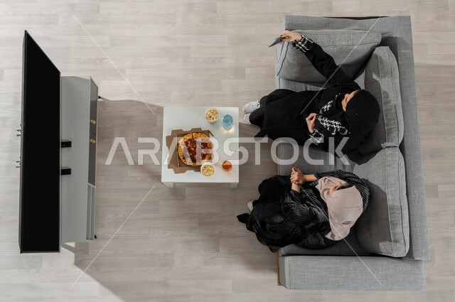 قضاء وقت الغداء في المنزل مع الأصدقاء، بورتريه علوي عامودي لفتاتان عربيتان خليجيتان سعوديتان ترتديان الحجاب والعباءة السوداء تجلسان على الأريكة في غرفة المعيشة وتقومان بتناول البيتزا، عرض شاشة الجوال واللابتوب السوداء الفارغة، خلفية بيج