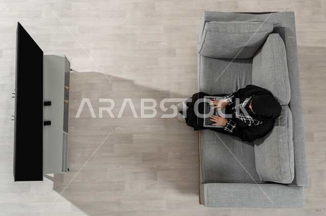 Using electronic devices, spending time at home, a vertical overhead portrait of a Saudi Gulf Arab girl wearing the hijab and black abaya, sitting on the sofa in the living room and holding a laptop in her hand, showing the empty black TV and laptop screen, beige background