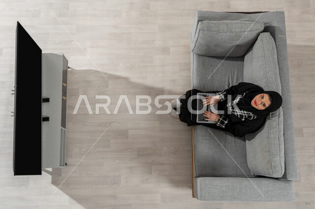 قضاء الوقت في المنزل، بورتريه علوي عامودي لفتاة عربية خليجية سعودية ترتدي الحجاب والعباءة السوداء تنظر إلى الكاميرا وتجلس على الأريكة في غرفة المعيشة وتقوم باستخدام جهاز الحاسوب المحمول، عرض شاشة التلفاز واللابتوب السوداء الفارغة، خلفية بيج