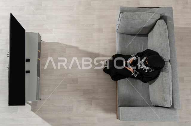 استخدام الأجهزة الذكية، قضاء الوقت في المنزل، بورتريه علوي عامودي لفتاة عربية خليجية سعودية ترتدي الحجاب والعباءة السوداء تجلس على الأريكة في غرفة المعيشة وتقول باستعمال  الهاتف المحمول، عرض شاشة التلفاز والجوال السوداء الفارغة، خلفية بيج