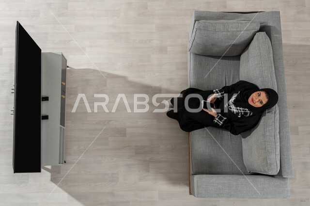 قضاء الوقت في المنزل، بورتريه علوي عامودي لفتاة عربية خليجية سعودية ترتدي الحجاب والعباءة السوداء تنظر إلى الكاميرا وتجلس على الأريكة في غرفة المعيشة وتقوم باستخدام الهاتف المحمول، عرض شاشة التلفاز والجوال السوداء الفارغة، خلفية بيج