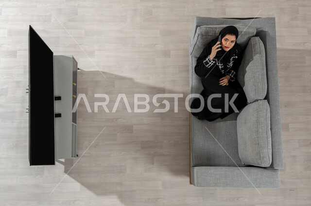 إجراء مكالمة هاتفية عبر الجوال، بورتريه علوي عامودي لفتاة عربية خليجية سعودية ترتدي الحجاب والعباءة السوداء تنظر إلى الكاميرا وتجلس على الأريكة في غرفة المعيشة وتقوم بالتحدث عبر الهاتف المحمول، عرض شاشة التلفاز السوداء الفارغة، خلفية بيج