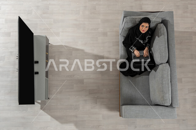 قضاء الوقت بإنجاز المهام المطلوبة،  بورتريه علوي عامودي لفتاة عربية خليجية سعودية مبتسمة ترتدي الحجاب والعباءة السوداء تنظر إلى الكاميرا وتجلس على الأريكة في غرفة المعيشة وتقوم بالعمل من خلال جهاز الحاسوب المحمول، عرض شاشة التلفاز السوداء الفارغة، خلفية بيج