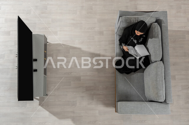 قضاء الوقت بالمطالعة والثقافة، ممارسة الهوايات المفضلة، بورتريه علوي عامودي لفتاة عربية خليجية سعودية ترتدي الحجاب والعباءة السوداء تجلس على الأريكة في غرفة المعيشة وتقوم بقراءة كتاب، عرض شاشة التلفاز السوداء الفارغة، خلفية بيج