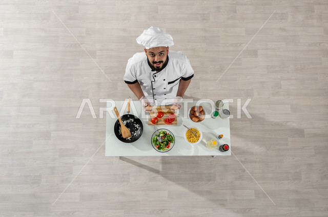 تجهيز وجبة صحية، فنون ومهارات الطبخ، بورتريه علوي عامودي لشيف عربي خليجي سعودي مبتسم يرتدي الزي المخصص للطهاة ينظر إلى الكاميرا ويقوم بإعداد الطعام وأمامه المكونات والمقادير المناسبة، اكتساب مهارات جديدة، خلفية بيج
