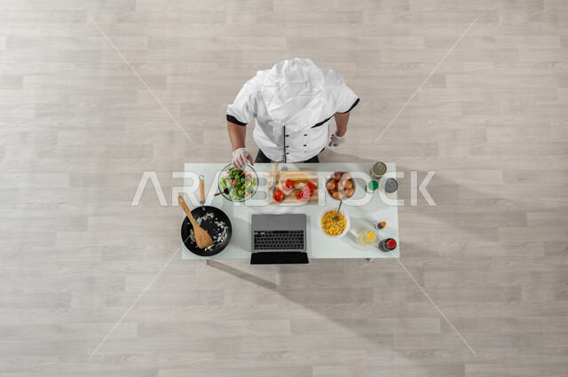 اكتساب مهارات جديدة أونلاين، تعلم وتطبيق فنون الطهي عبر المواقع الإلكترونية، بورتريه علوي عامودي لشيف عربي خليجي سعودي يرتدي الزي المخصص للطهاة وأمامه المكونات والمقادير المناسبة، تصوير وعرض الطبخ من خلال جهازالحاسوب المحمول، خلفية بيج