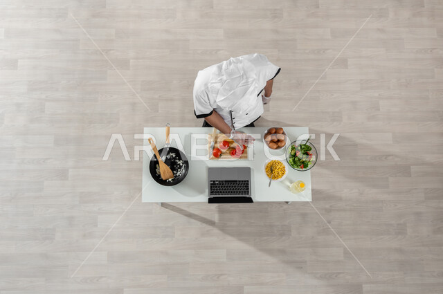 تعلم وتطبيق فنون الطهي عبر المواقع الإلكترونية، بورتريه علوي عامودي لشيف عربي خليجي سعودي يرتدي الزي المخصص للطهاة وأمامه المكونات والمقادير المناسبة، تصوير وعرض الطبخ من خلال جهازالحاسوب المحمول، تجهيز وتحضير وجبة طعام صحية، اكتساب مهارات جديدة أونلاين، خلفية بيج