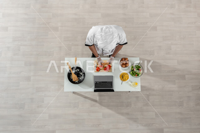 تجهيز وتحضير وجبة طعام صحية، تعلم وتطبيق فنون الطهي عبر المواقع الإلكترونية، بورتريه علوي عامودي لشيف عربي خليجي سعودي يرتدي الزي المخصص للطهاة وأمامه المكونات والمقادير المناسبة، تصوير وعرض الطبخ من خلال جهازالحاسوب المحمول، اكتساب مهارات جديدة أونلاين، خلفية بيج