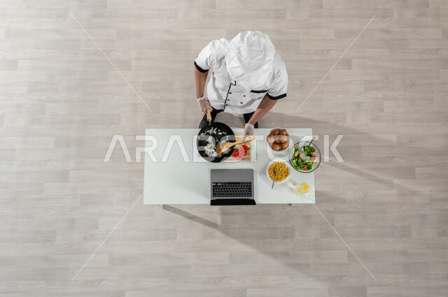 تعلم وتطبيق فنون الطهي من خلال المواقع الإلكترونية، بورتريه علوي عامودي لشيف عربي خليجي سعودي يرتدي الزي المخصص للطهاة يقوم بتقليب شرائح البصل وأمامه المكونات والمقادير المناسبة، اكتساب مهارات جديدة أونلاين، تصوير وعرض الطبخ عبر الحاسوب المحمول، خلفية بيج