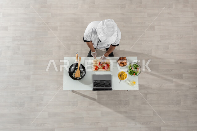 تعلم وتطبيق  فنون الطهي من خلال المواقع الإلكترونية، بورتريه علوي عامودي لشيف عربي خليجي سعودي يرتدي الزي المخصص للطهاة يقوم بتقطيع الطماطم إلى شرائح وأمامه المكونات والمقادير المناسبة، تصوير وعرض الطبخ عبر الحاسوب المحمول، خلفية بيج