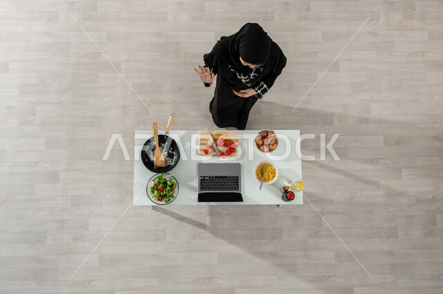 تصوير وعرض الطبخ عبر جهاز الحاسوب المحمول، بورتريه علوي عامودي لفتاة عربية خليجية سعودية ترتدي الحجاب والعباءة السوداء تقوم بإيماءات الانتهاء والسلام وأمامها المكونات والمقادير المناسبة، تعلم وتطبيق فنون الطهي من خلال المواقع الإلكترونية، خلفية بيج