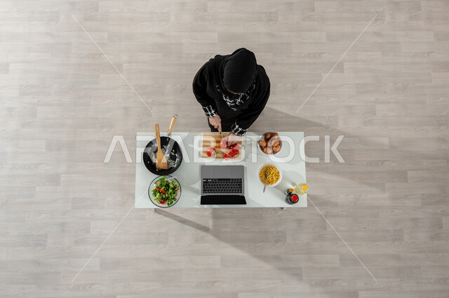 اكتساب مهارات جديدة أونلاين، تعلم وتطبيق فنون الطهي من خلال المواقع الإلكترونية، بورتريه علوي عامودي لفتاة عربية خليجية سعودية ترتدي الحجاب والعباءة السوداء تقوم بتقطيع الطماطم إلى شرائح وأمامها المكونات والمقادير المناسبة، تصوير وعرض الطبخ عبر جهاز الحاسوب المحمول، خلفية بيج