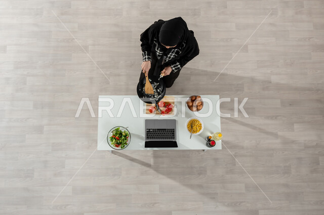 تعلم وتطبيق فنون الطهي من خلال المواقع الإلكترونية، بورتريه علوي عامودي لفتاة عربية خليجية سعودية ترتدي الحجاب والعباءة السوداء تقوم بتحميص شرائح البصل وأمامها المكونات والمقادير المناسبة، تصوير وعرض الطبخ عبر جهاز الحاسوب المحمول، اكتساب مهارات جديدة أونلاين، خلفية بيج