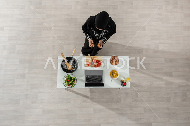 تعلم وتطبيق فنون الطهي من خلال المواقع الإلكترونية، بورتريه علوي عامودي لفتاة عربية خليجية سعودية ترتدي الحجاب والعباءة السوداء تقوم بتصوير وعرض الطبخ عبر جهاز اللابتوب وأمامها المكونات والمقادير اللازمة، اكتساب مهارات جديدة أونلاين، شرح المكونات والكميات المطلوبة، خلفية بيج