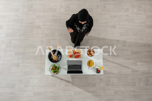 التفاعل مع المتابعين بإيماءات الترحيب من خلال البث المباشر عبر المواقع الإلكترونية، بورتريه علوي عامودي لفتاة عربية خليجية سعودية ترتدي الحجاب والعباءة السوداء تقوم بتصوير وعرض الطبخ عبر جهاز الحاسوب المحمول وأمامها المكونات والمقادير اللازمة، خلفية بيج