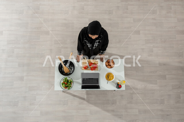 تعلم وتطبيق فنون الطهي من خلال المواقع الإلكترونية، بورتريه علوي عامودي لفتاة عربية خليجية سعودية ترتدي الحجاب والعباءة السوداء تقوم بتصوير وعرض الطبخ عبر جهاز الحاسوب المحمول وأمامها المكونات والمقادير اللازمة، اكتساب مهارات جديدة أونلاين، خلفية بيج
