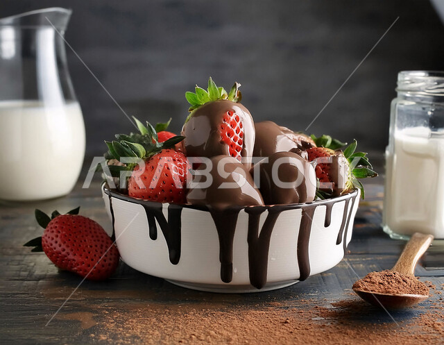 Ripe pieces of delicious strawberry fruit with chocolate in a white bowl on a wooden surface, a healthy milk drink in a glass bowl, local Saudi agricultural crops and products, fresh seasonal fruits, black background