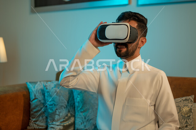 قضاء اوقات ممتعة في المنزل، استخدام الأجهزة والتقنيات الحديثة، إيماءات الاندماج والتفاعل مع الألعاب الافتراضية، الاستمتاع بمشاهدة أفلام العالم الخيالي، رجل عربي خليجي سعودي يرتدي الثوب التقليدي يضع نظارة الواقع الافتراضي ثلاثية الابعاد