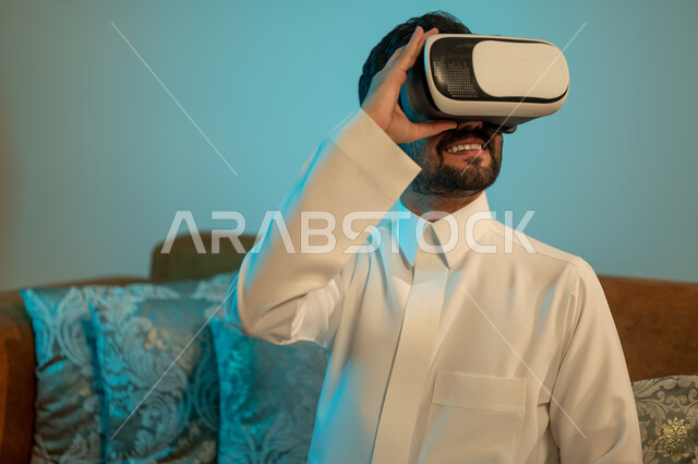 إيماءات الاندماج والتفاعل مع الألعاب الافتراضية، الاستمتاع بمشاهدة أفلام العالم الخيالي، رجل عربي خليجي سعودي يرتدي الثوب التقليدي يضع نظارة الواقع الافتراضي ثلاثية الابعاد، قضاء اوقات ممتعة في المنزل، استخدام الأجهزة والتقنيات الحديثة