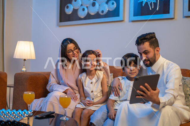 ايماءات السعادة والسرور، متابعة الافلام والمسلسلات باستخدام التابلت، اجراء مكالمات فيديو مع الاقارب،  أجواء اسرية سعيدة في المنزل، زوجان عربيان خلجيان سعوديان يقومان بشرب العصير برفقه ابنائهما، قضاء اوقات عائلية ممتعة
