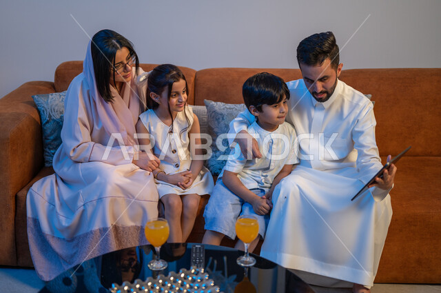 Making video calls with relatives, expressions of happiness and joy, a happy family atmosphere at home, a Saudi Gulf Arab couple drinking juice with their children, spending enjoyable family times, watching movies and series using the tablet.